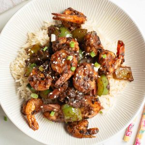 A bowl of white rice topped with shrimp, green bell peppers, and a dark sauce, garnished with sesame seeds and chopped green onions.