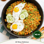 A skillet of Indonesian fried rice (nasi goreng) with vegetables, two fried eggs, and cucumber slices, on a table with garnish bowls and a green-and-white cloth nearby.
