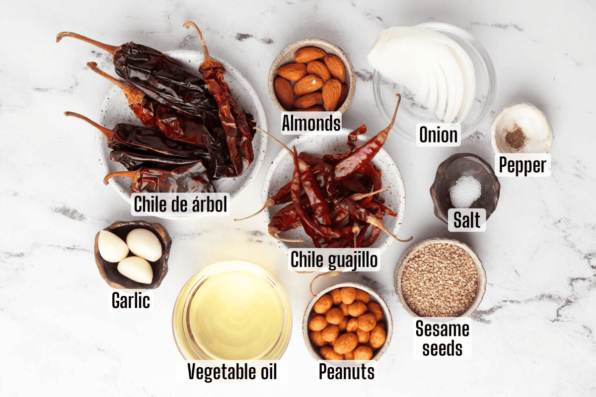 Labeled ingredients for a recipe on a white surface, including dried chiles, almonds, peanuts, sesame seeds, onion, garlic, vegetable oil, salt, and pepper.