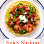 A tostada topped with mashed avocado, spicy shrimp, tomato, onion, jalapeño slices, cilantro, and a dollop of sour cream on a white plate.