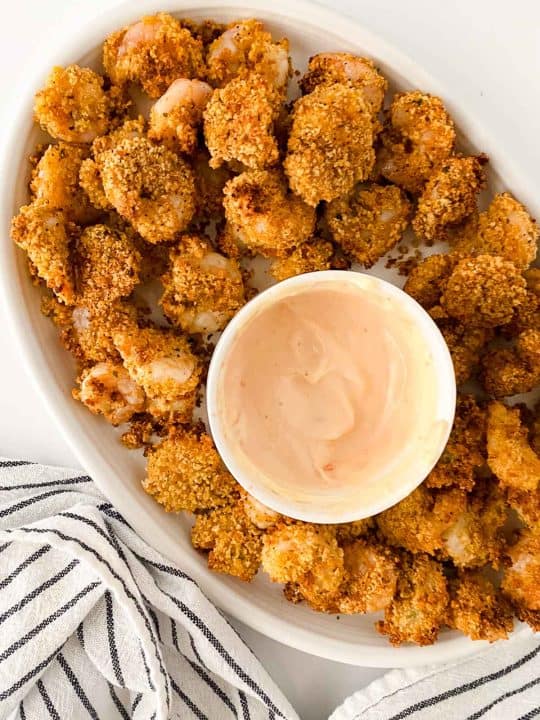 A plate of breaded, baked shrimp arranged around a bowl of creamy dipping sauce, with a striped cloth napkin beside the plate.