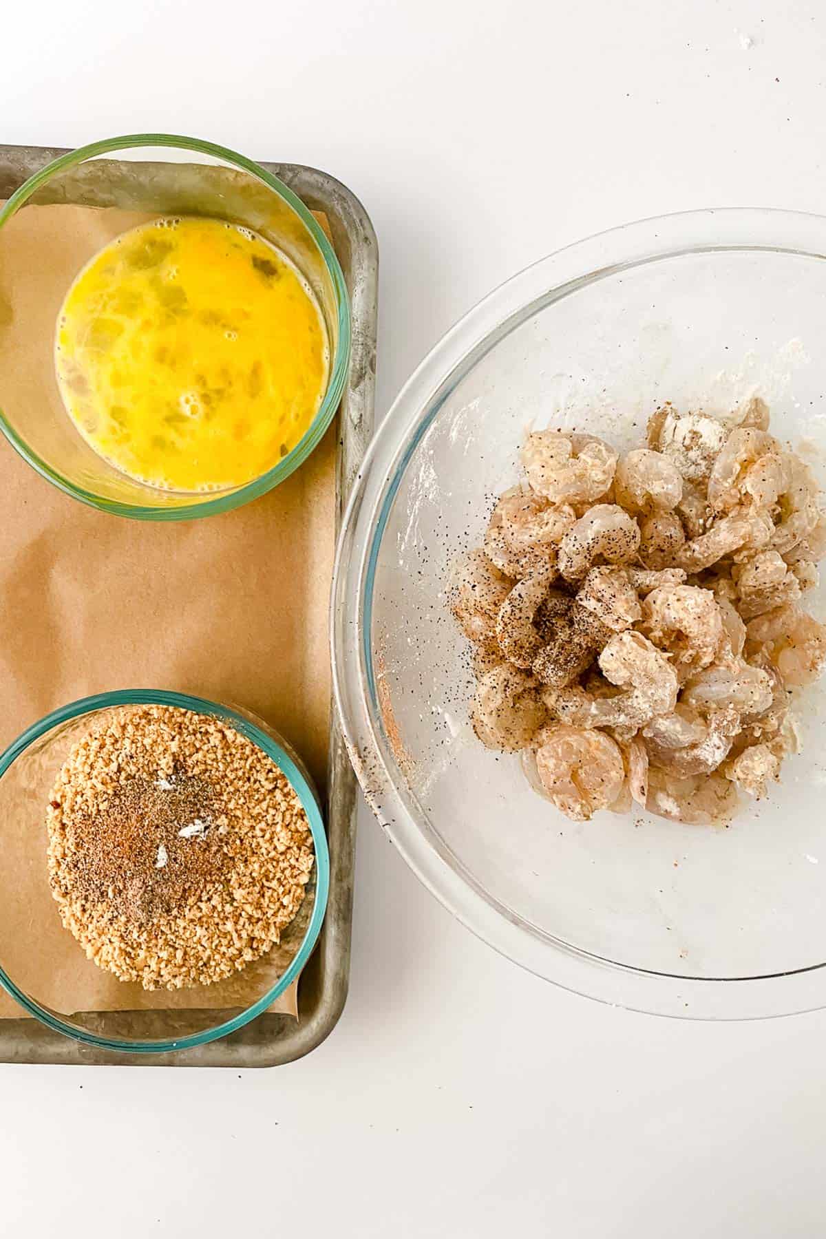 Three bowls on a tray: one with beaten eggs, one with seasoned breadcrumbs, and one with raw seasoned shrimp.