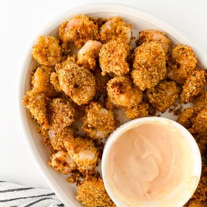 A plate of breaded, baked shrimp is served with a small bowl of creamy dipping sauce on the side.