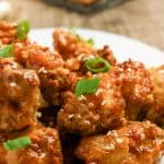 Close-up of fried chicken pieces glazed with sauce, garnished with sesame seeds and chopped green onions on a white plate.