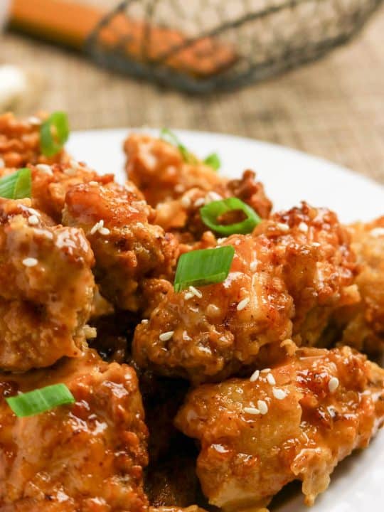 Close-up of fried chicken pieces glazed with sauce, garnished with sesame seeds and chopped green onions on a white plate.