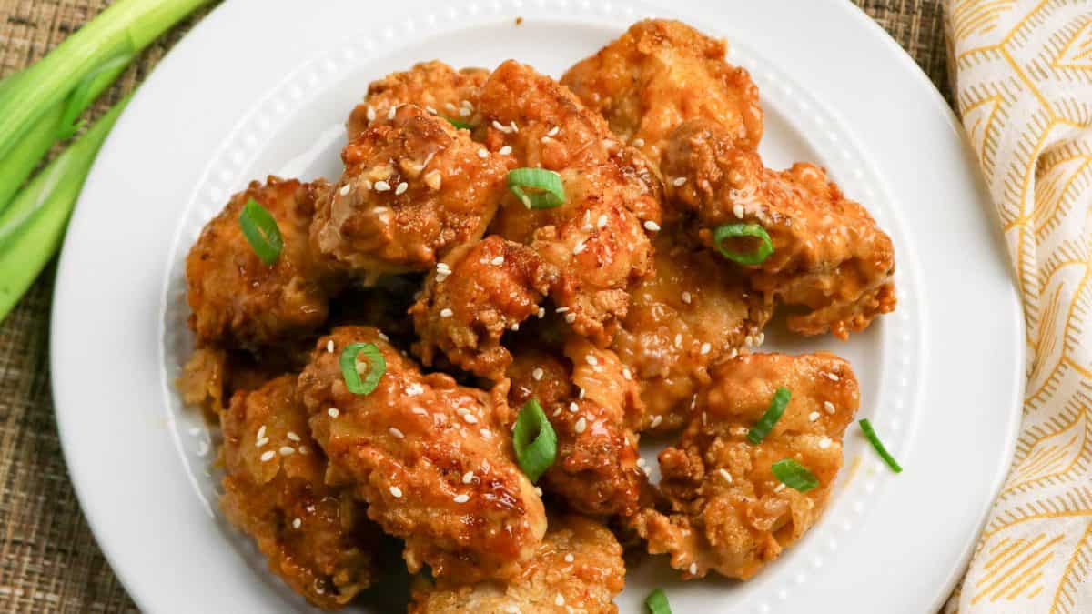 A white plate of fried chicken pieces garnished with sesame seeds and sliced green onions, placed on a woven surface next to a yellow-patterned napkin.