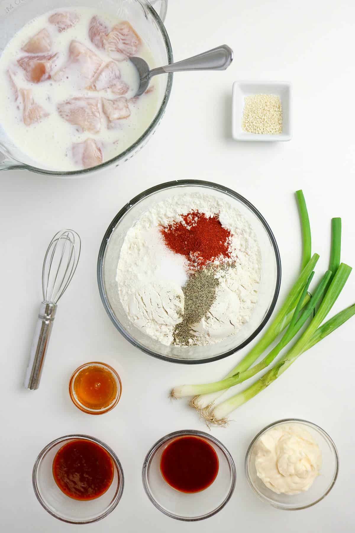 Bowls of chicken in buttermilk, flour with spices, sesame seeds, scallions, whisk, and various sauces are arranged on a white surface for cooking preparation.