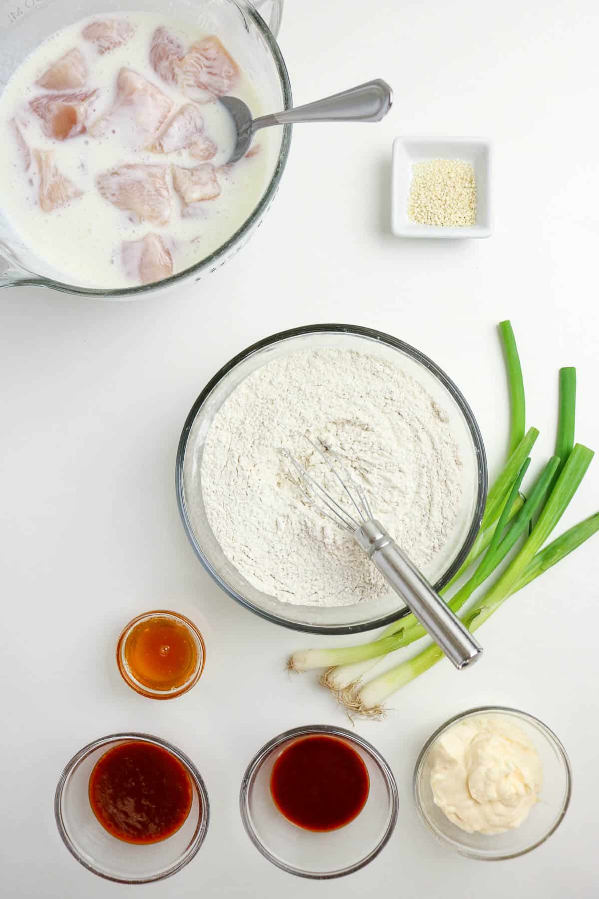 Bowls of raw chicken in buttermilk, flour with a whisk, sesame seeds, three sauces, mayonnaise, and green onions arranged on a white surface.