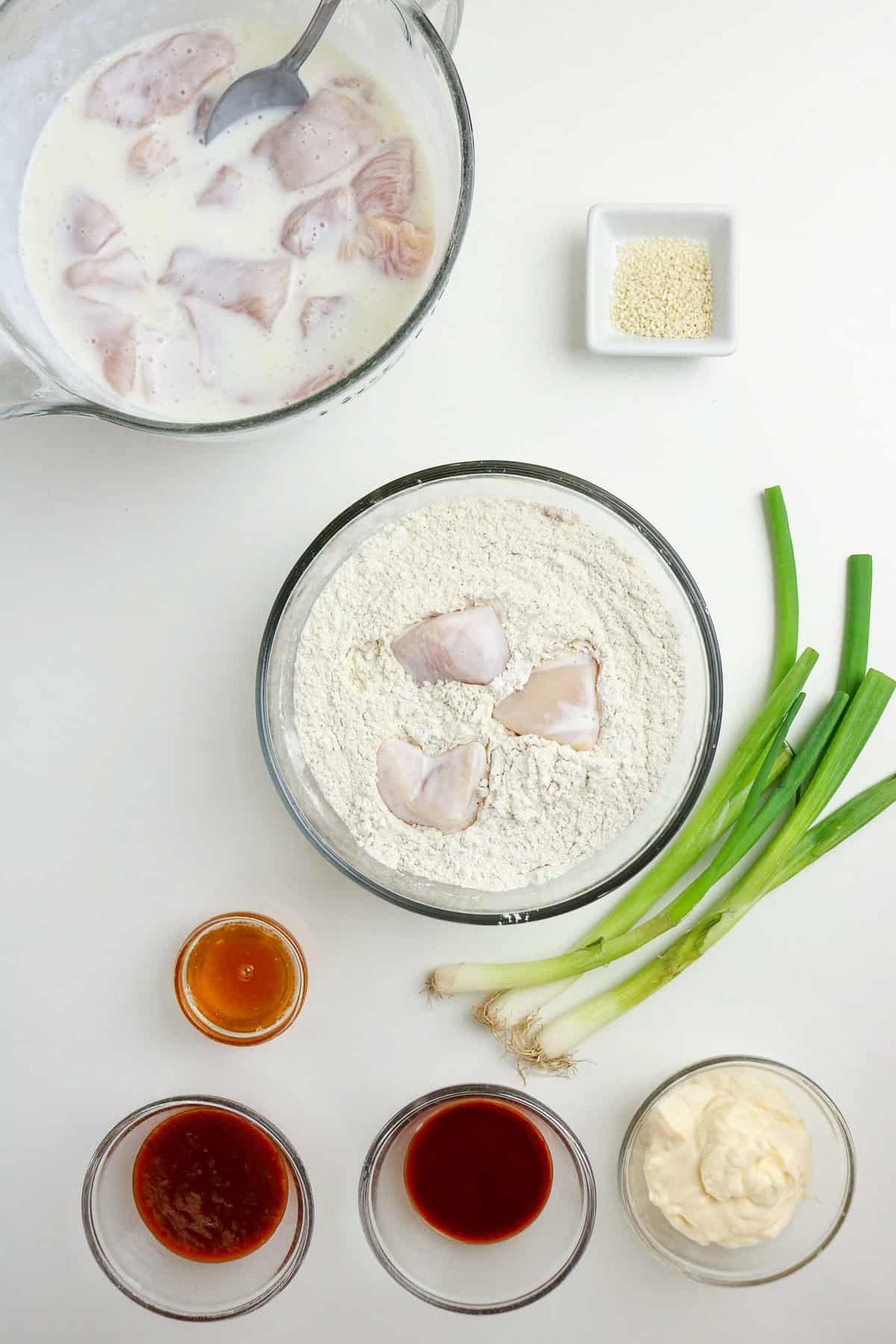 Raw chicken pieces in a milk mixture, chicken in flour, green onions, sesame seeds, and four sauces in small bowls are arranged on a white surface.