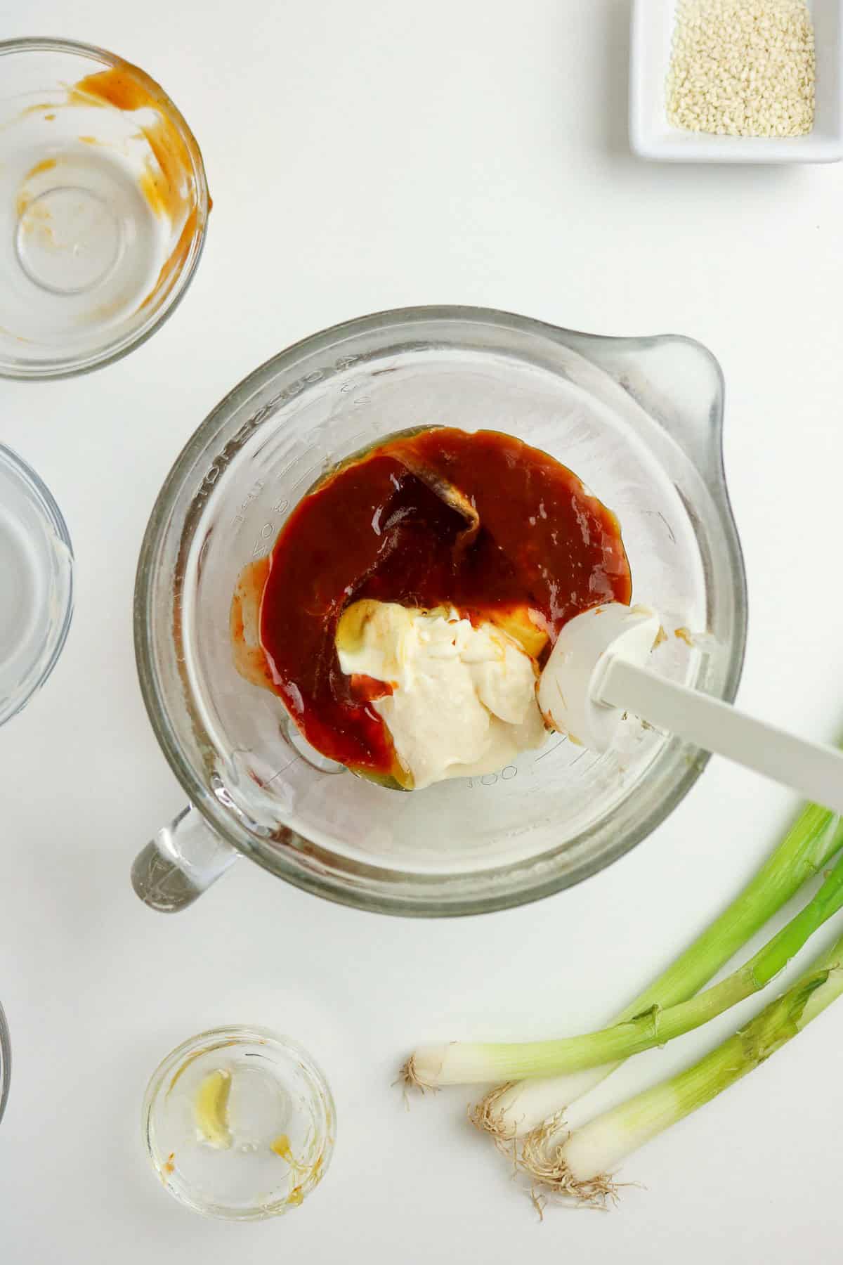Glass mixing bowl with mayonnaise, brown sauce, and spoon on a white surface, surrounded by empty bowls, sesame seeds, and two green onions.