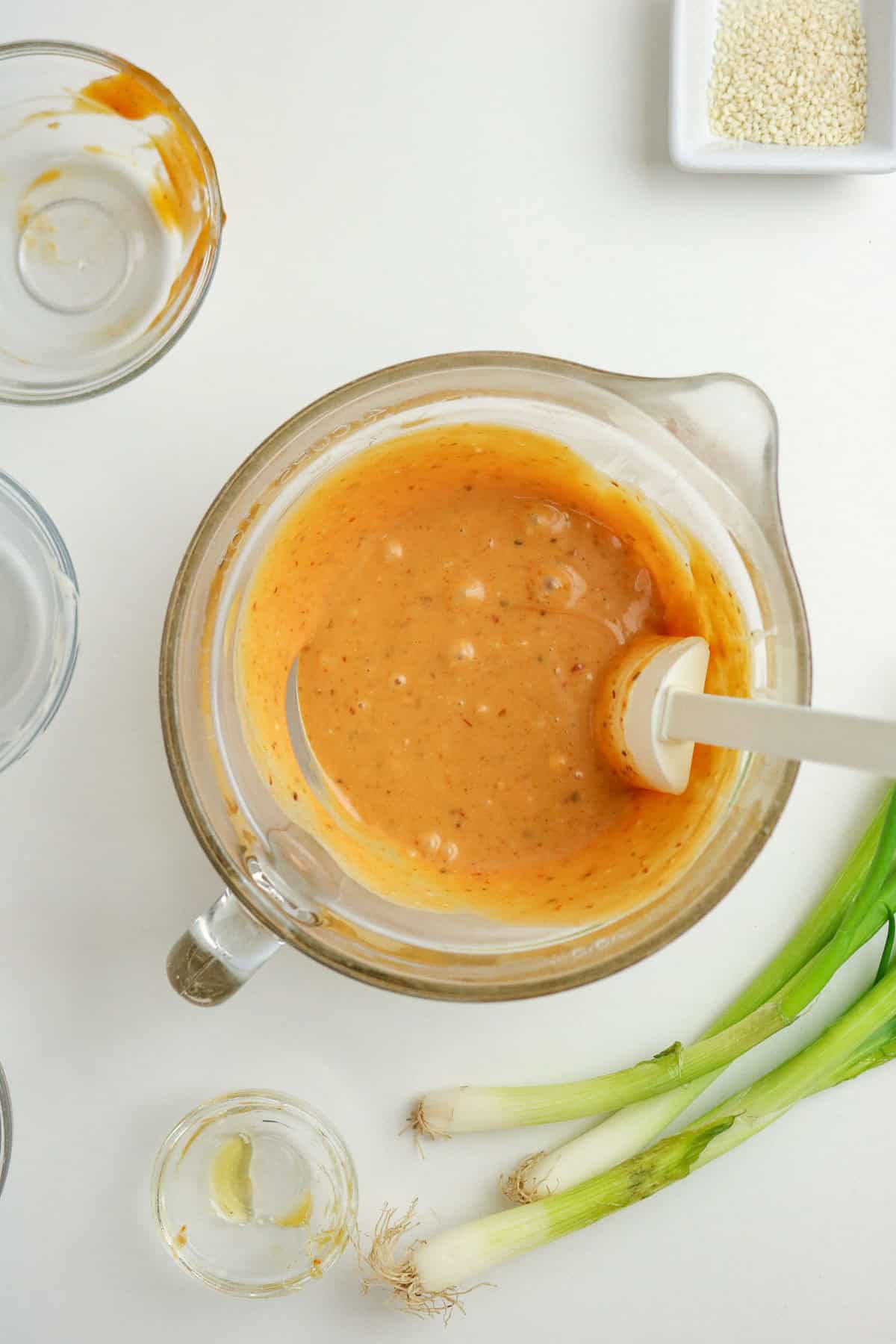 A glass bowl with creamy orange sauce and a spatula, surrounded by green onions, an empty glass, and a dish of sesame seeds on a white surface.