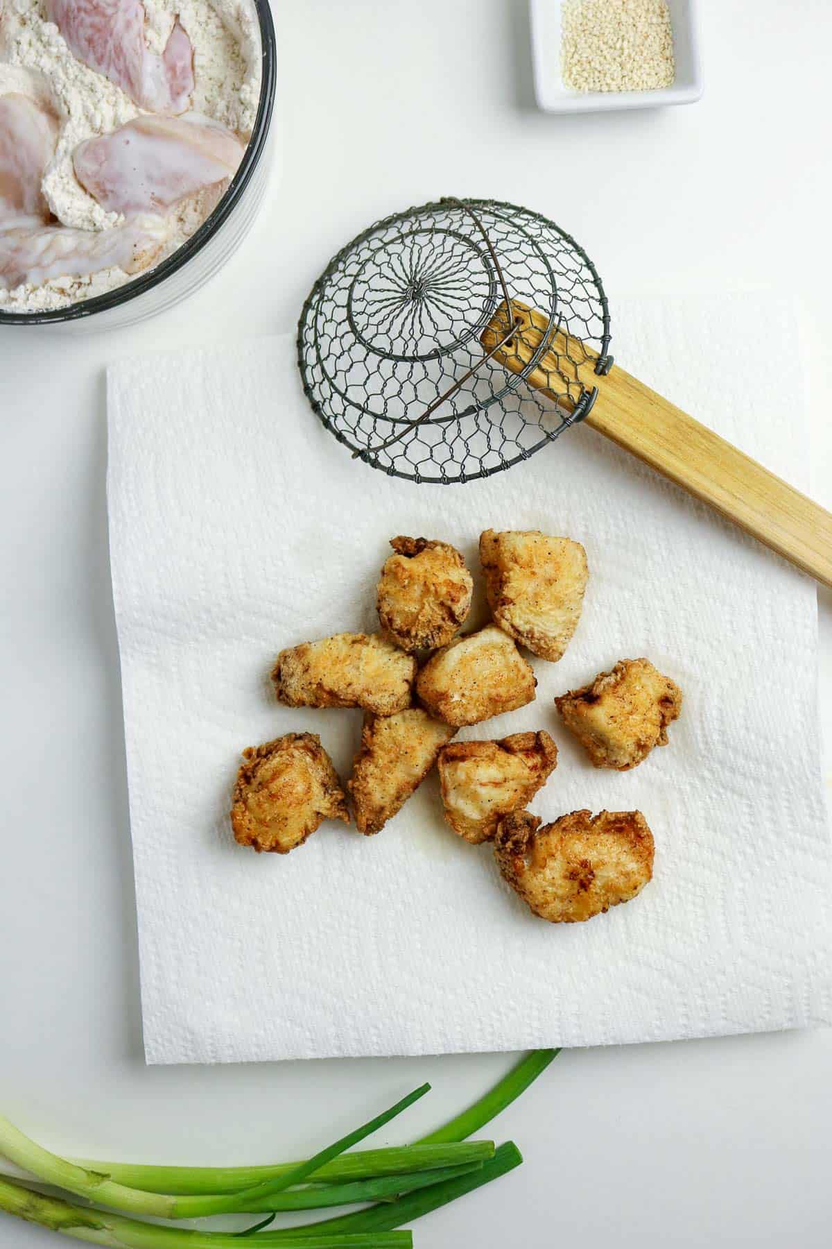 Seven pieces of fried chicken sit on a paper towel near a wire skimmer, a bowl of raw chicken in flour, a small dish of sesame seeds, and green onions on a white surface.
