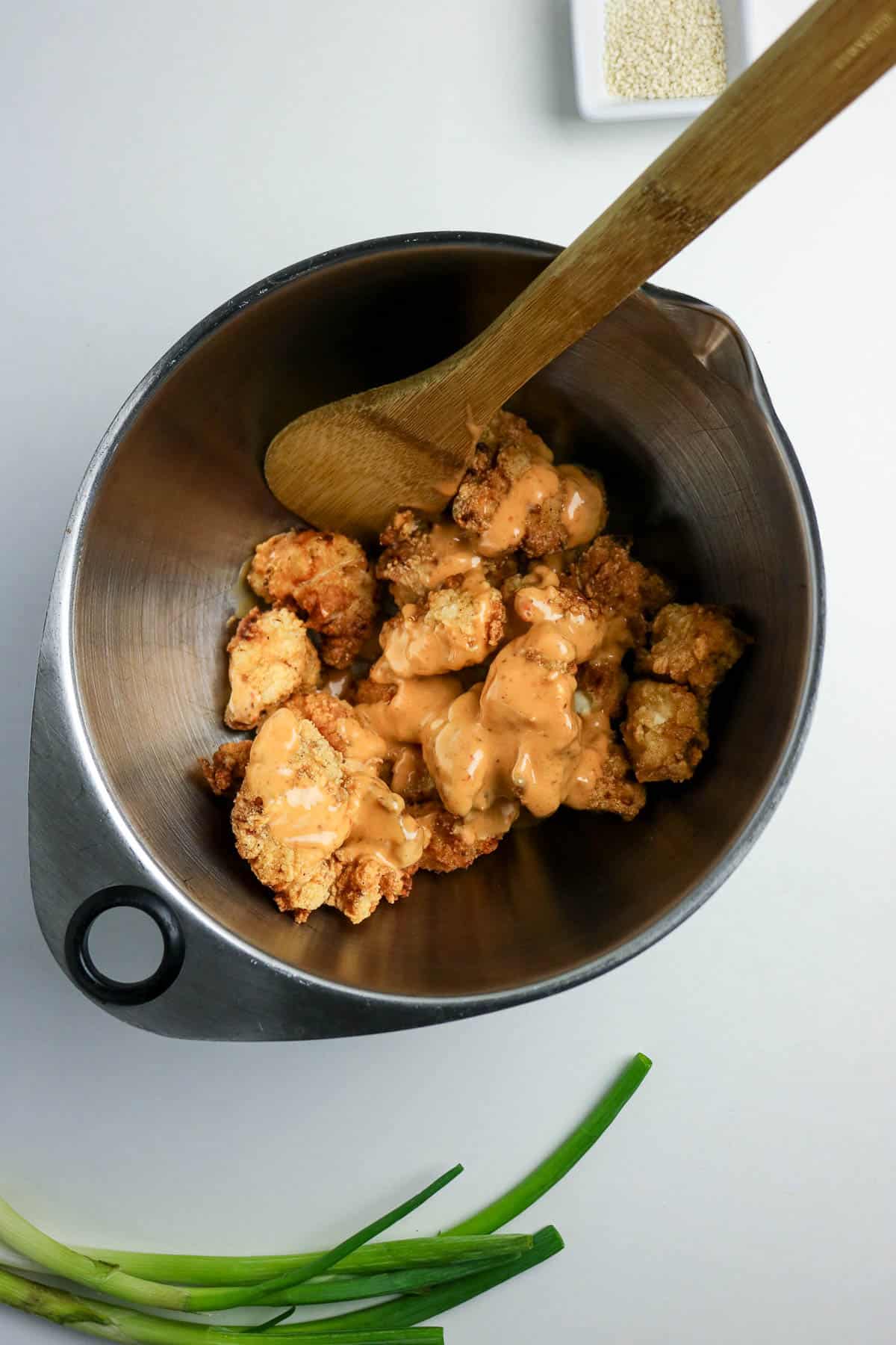 A metal mixing bowl with fried chicken pieces and creamy sauce, stirred with a wooden spoon. Green onions and a dish of sesame seeds are nearby.