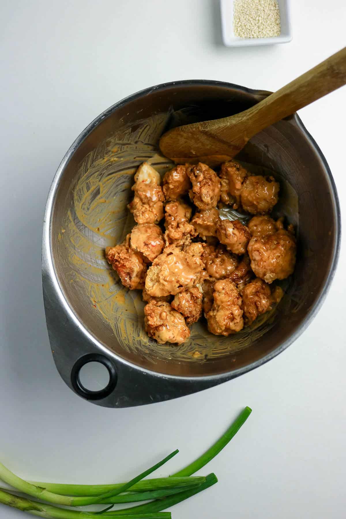 Metal bowl with sauced chicken pieces and a wooden spoon; green onions and a small dish of sesame seeds are nearby on a white surface.