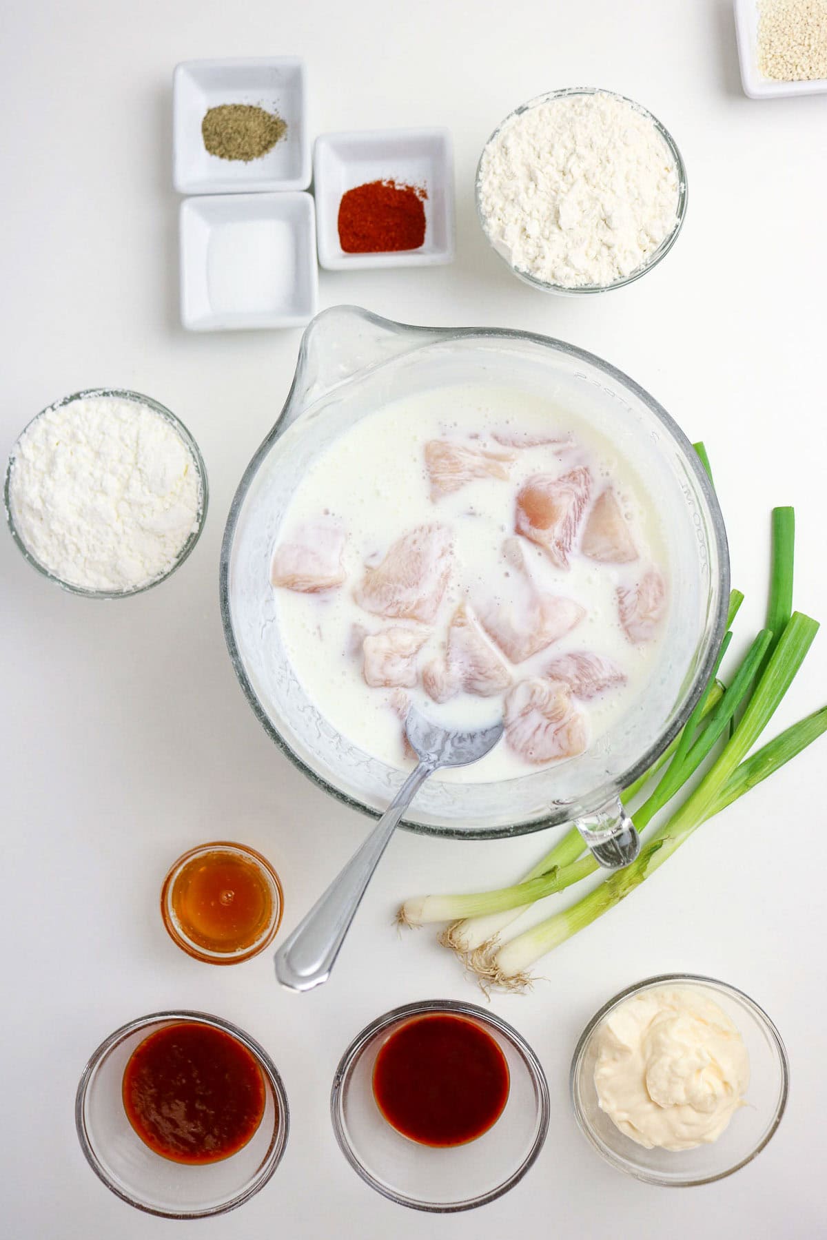 Raw fish pieces in a bowl of milk with a spoon, surrounded by flour, seasonings, sauces, mayonnaise, and green onions, arranged on a white surface.