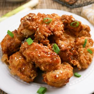A white plate with pieces of fried chicken coated in sauce, sprinkled with sesame seeds and garnished with chopped green onions.