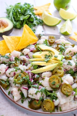 A plate of ceviche topped with sliced avocado, jalapeños, cilantro, and red onion, served with tortilla chips. Lime wedges, cilantro, and half an avocado are in the background.