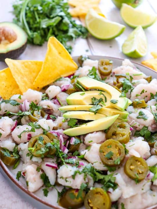 A plate of ceviche topped with sliced avocado, jalapeños, cilantro, and red onion, served with tortilla chips. Lime wedges, cilantro, and half an avocado are in the background.