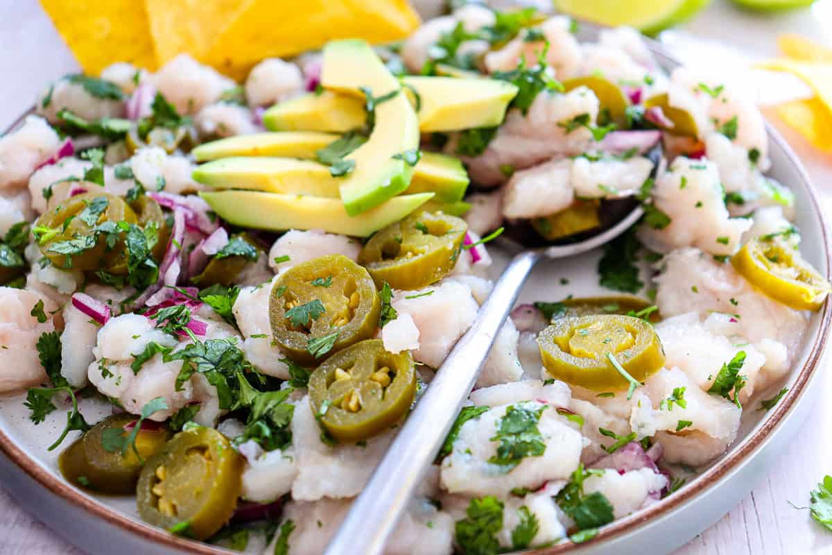 A plate of ceviche garnished with sliced avocado, pickled jalapeños, cilantro, and red onions, with a spoon and tortilla chips on the side.