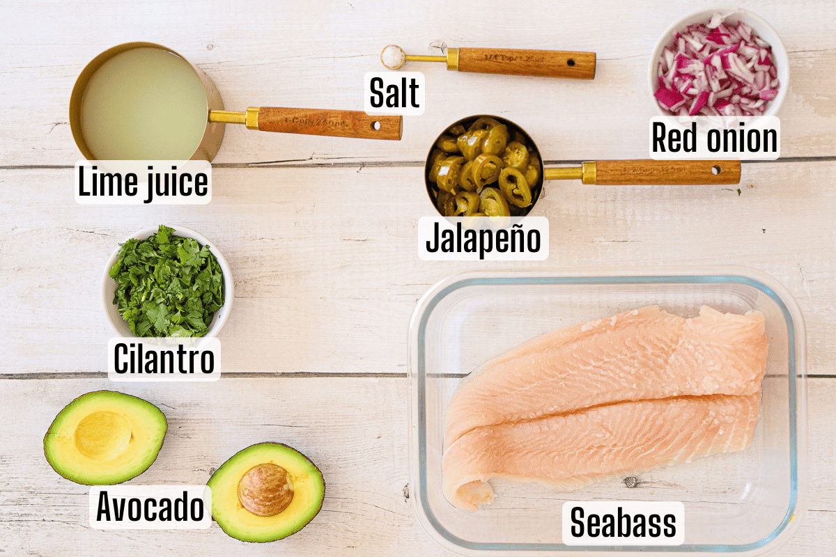 Ingredients for ceviche including lime juice, salt, jalapeño, red onion, cilantro, avocado, and raw seabass displayed on a white wooden surface.