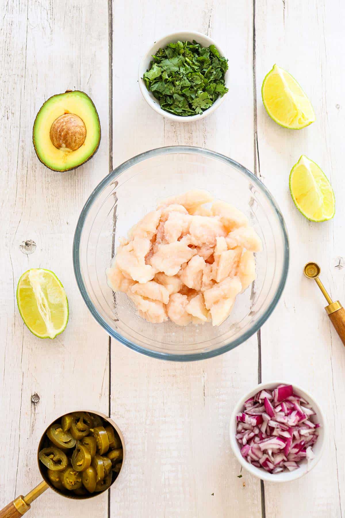 A glass bowl of raw shrimp surrounded by chopped cilantro, lime wedges, avocado, sliced jalapeños, and diced red onion on a white wooden surface.