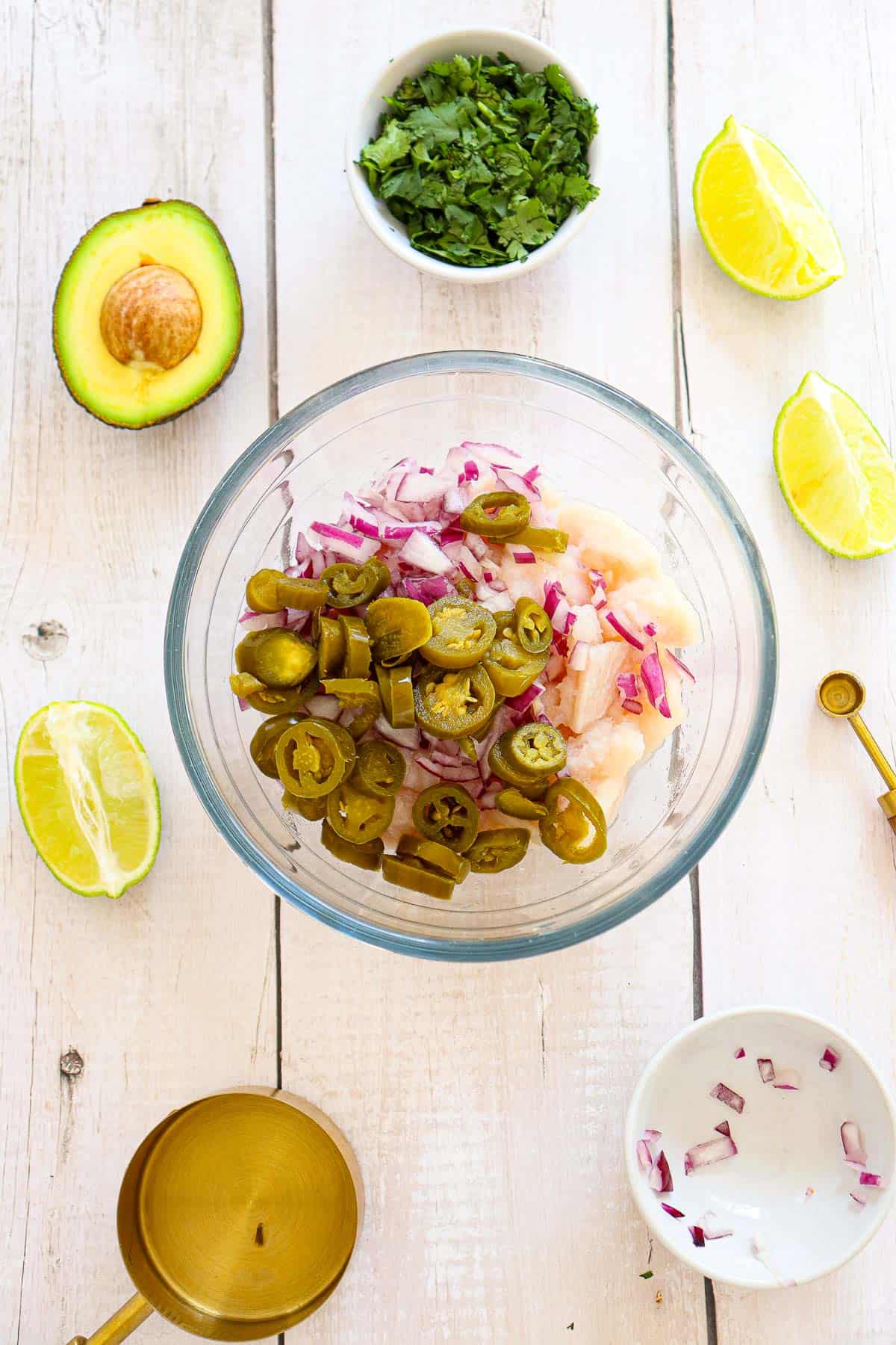 A glass bowl with chopped red onion, jalapeños, and raw chicken, surrounded by avocado, lime wedges, cilantro, and measuring spoons on a white wooden surface.