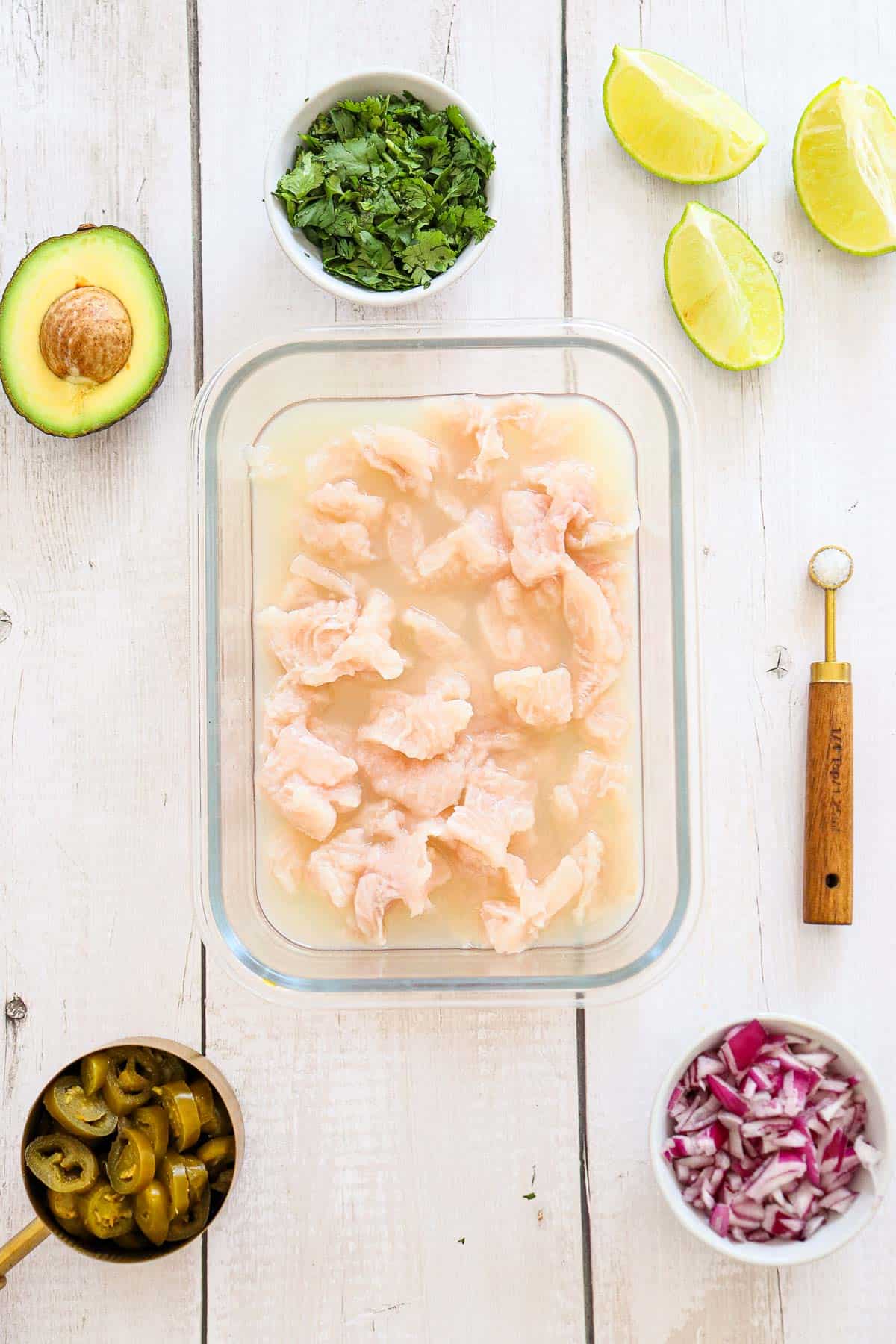Glass dish of raw fish marinating in liquid, surrounded by avocado, lime wedges, chopped cilantro, red onion, and sliced jalapeños on a white wooden surface.