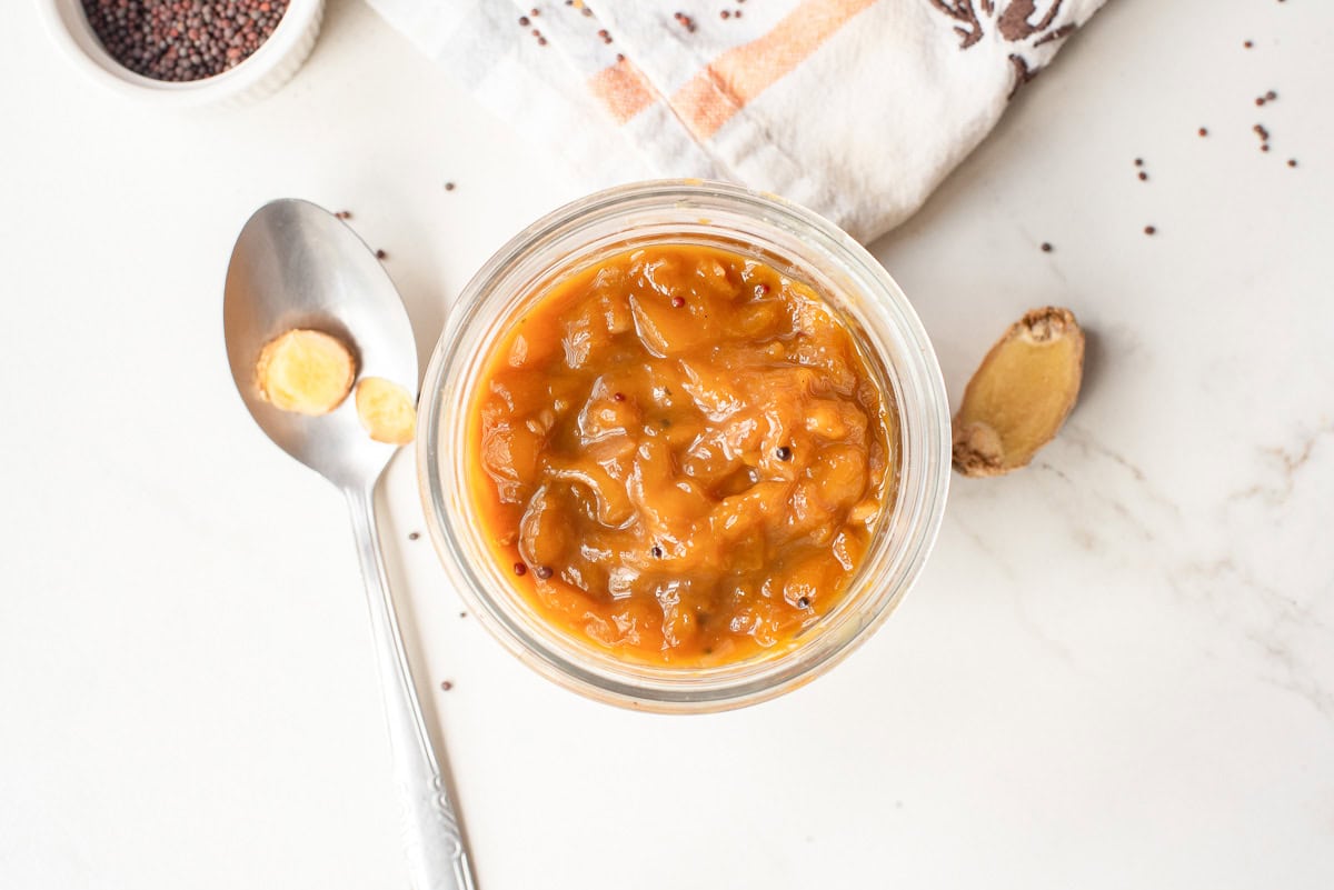 A glass jar of orange mango chutney sits on a white surface, next to a spoon with ginger slices, a ginger root, mustard seeds, and a folded napkin.