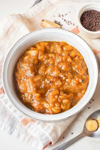 A white bowl filled with chunky, orange-brown mango chutney sits on a striped towel, with mustard seeds and a piece of ginger nearby.