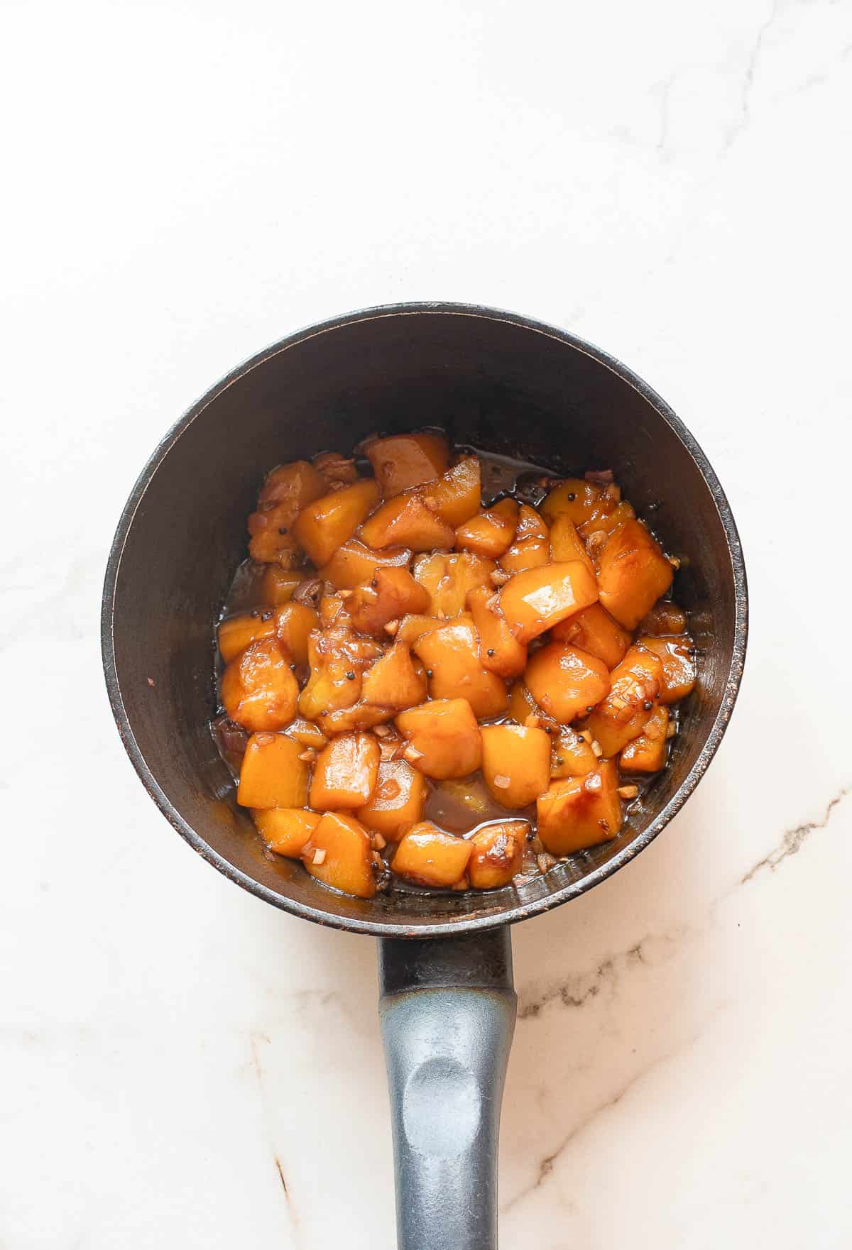A black saucepan filled with cooked, diced peaches in a syrupy sauce sits on a white marble surface.