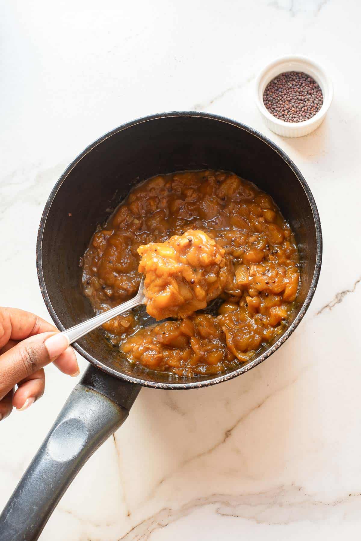 A hand holds a spoonful of thick, orange-brown chutney above a black saucepan, with a small bowl of mustard seeds nearby on a white marble surface.