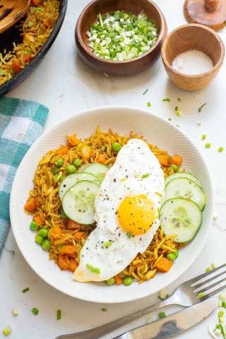 A plate of fried rice with peas, carrots, and sliced cucumber, topped with a sunny-side-up egg and black pepper, on a white table with bowls of chopped green onions and salt nearby.