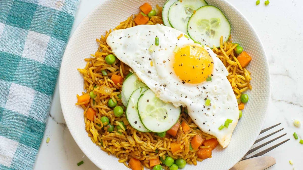 A plate of fried rice with peas, carrots, cucumber slices, and a sunny-side-up egg on top.