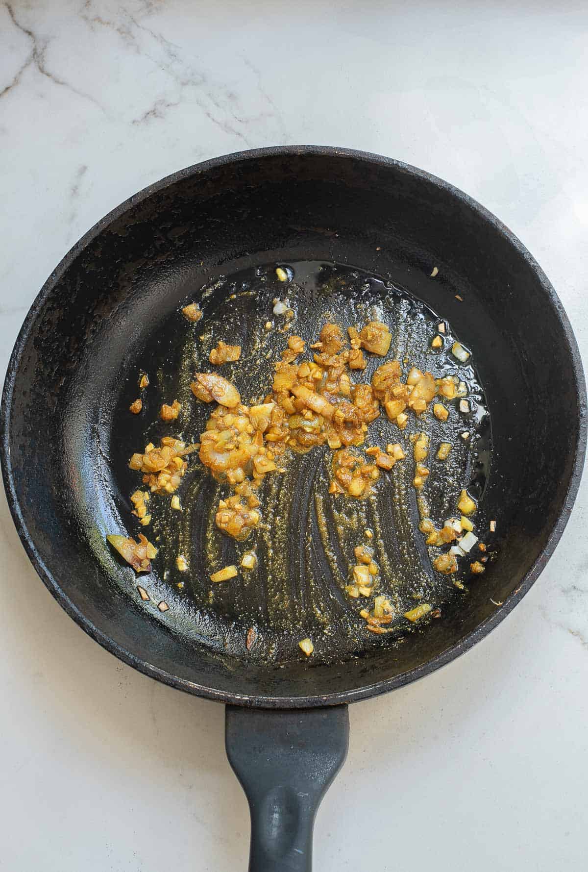 A black frying pan with sautéed chopped onions and garlic on a white marble surface.