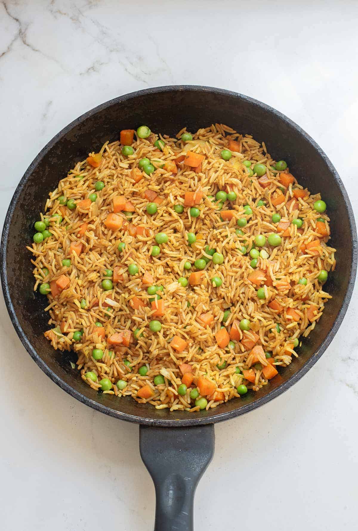 A black skillet containing cooked rice mixed with green peas and diced carrots, placed on a white marble surface.