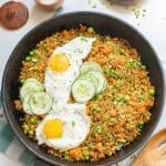 A skillet with fried rice, peas, carrots, two sunny side up eggs, and cucumber slices. Bowls of chopped green onions and salt, a wooden spoon, and a towel are nearby on a white surface.