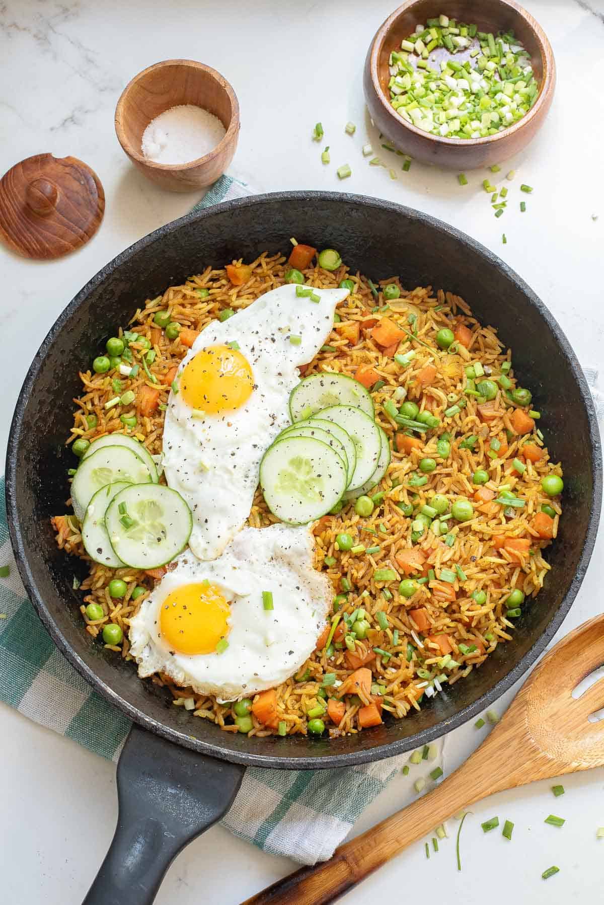 A skillet with fried rice, peas, carrots, two sunny side up eggs, and cucumber slices. Bowls of chopped green onions and salt, a wooden spoon, and a towel are nearby on a white surface.
