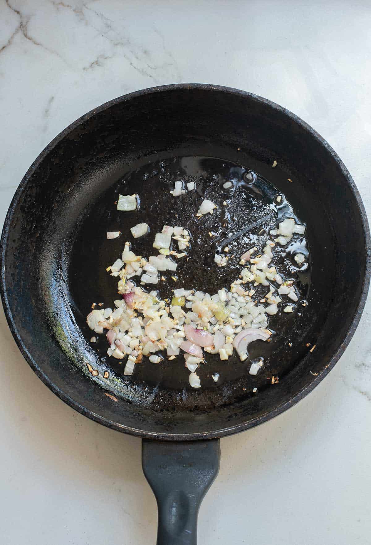 Chopped onions and garlic are sautéing in oil in a black frying pan on a white marble surface.