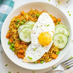 A white plate with fried rice, peas, carrots, three cucumber slices, and a sunny-side-up egg on top, garnished with chopped chives and black pepper.