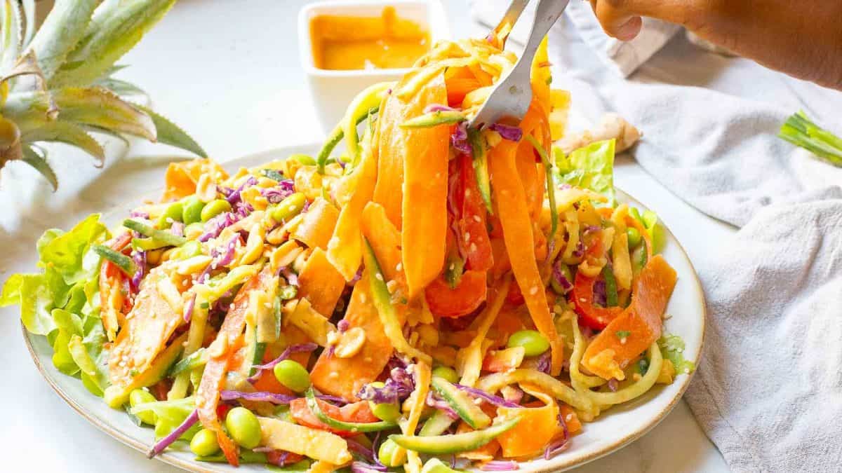 A fork lifts a portion of a colorful salad with spiralized vegetables, edamame, and lettuce on a white plate, with a small dish of dressing in the background.
