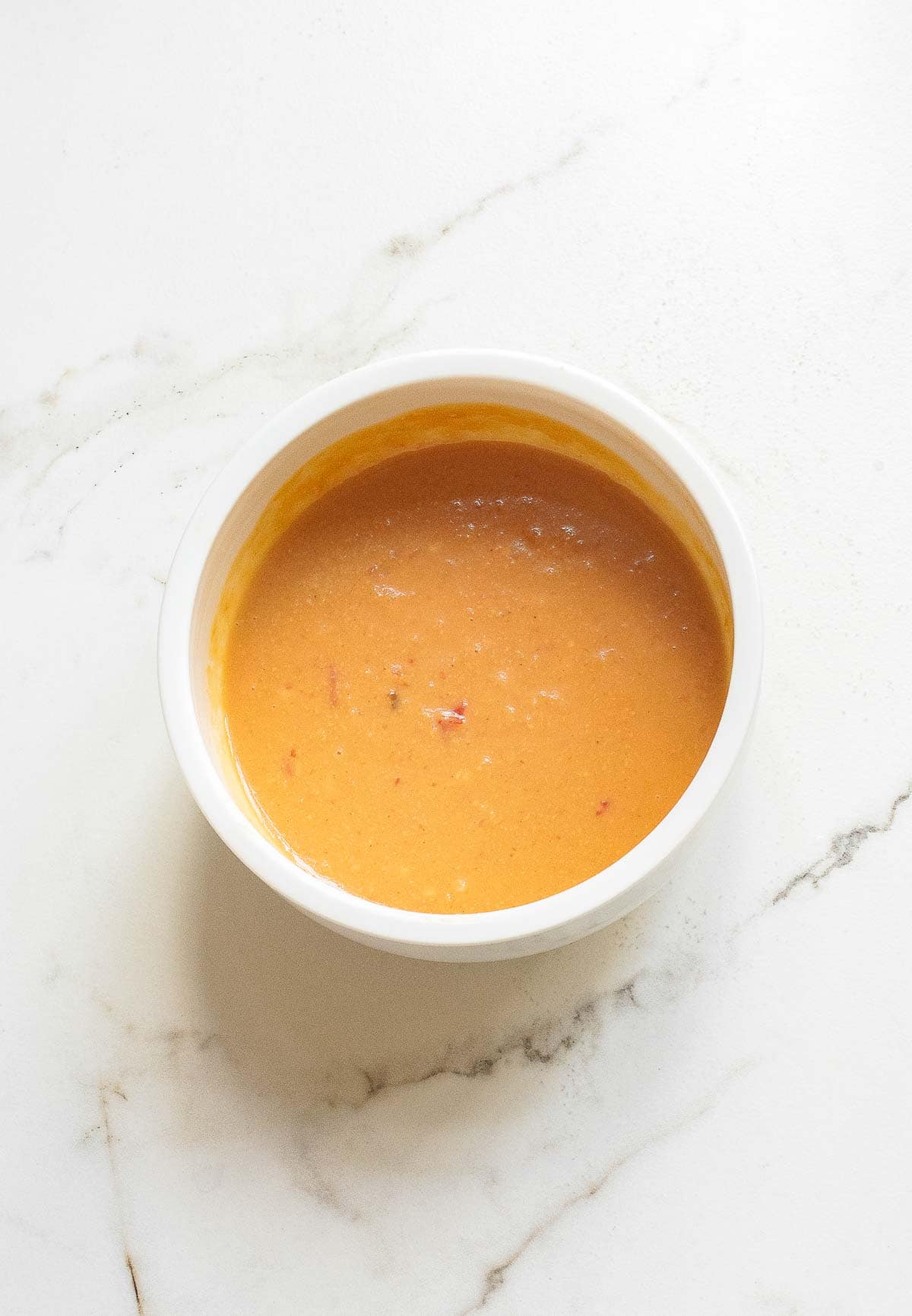 A white bowl filled with a smooth, orange-colored sauce sits on a white marble surface.