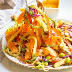 A plate of colorful spiralized vegetable salad with shredded carrots, cucumbers, cabbage, and edamame, being lifted with a fork and spoon. A dish of orange dressing is in the background.