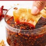 A hand dips a piece of tortilla chip into a clear glass bowl of vibrant salsa macha, its deep red color flecked with visible chili and seeds.