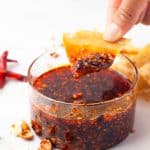 A hand dips a chip into a clear bowl of chili oil with visible chili flakes, surrounded by scattered chip pieces and garlic skins on a white surface.