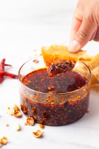 A hand dips a chip into a clear bowl of chili oil with visible chili flakes, surrounded by scattered chip pieces and garlic skins on a white surface.