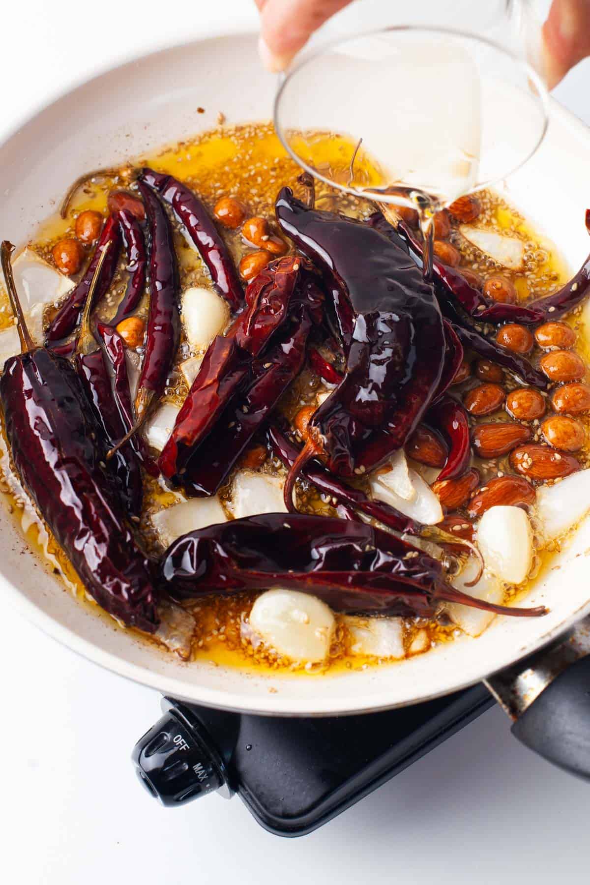 A skillet with dried chilies, garlic cloves, peanuts, and oil is being poured with clear liquid, likely for cooking or sautéing.