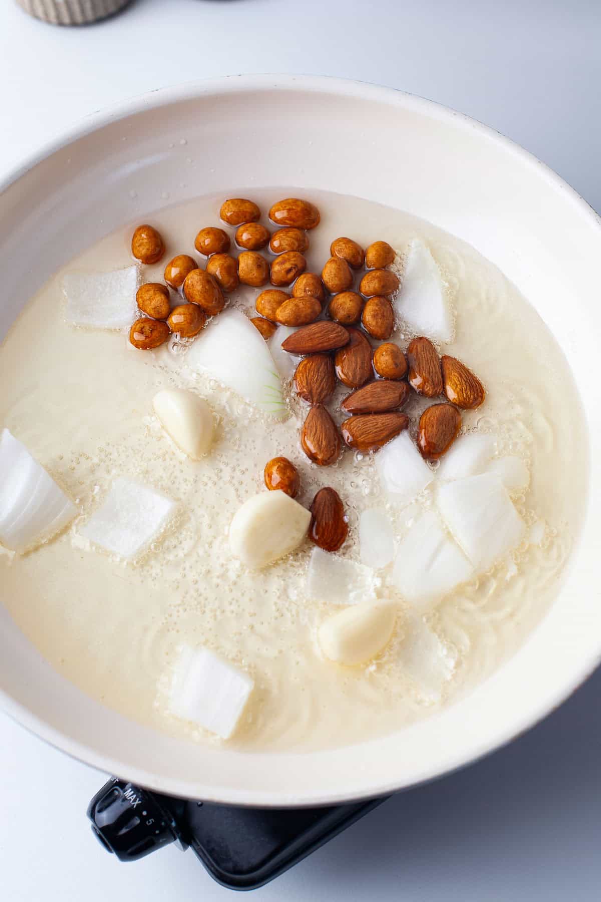 Raw almonds, whole garlic cloves, chopped onion, and peanuts are in oil, heating in a white pan on a stove.