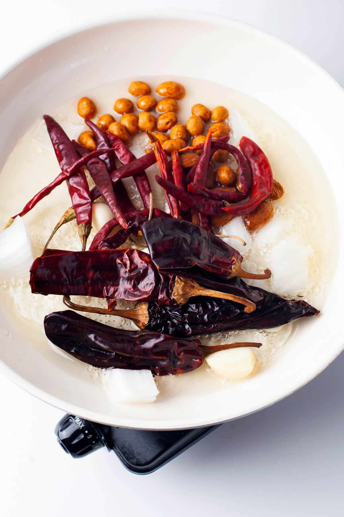 A white pan with dried red chilies, peanuts, onion chunks, garlic cloves, and oil heating on a stovetop.