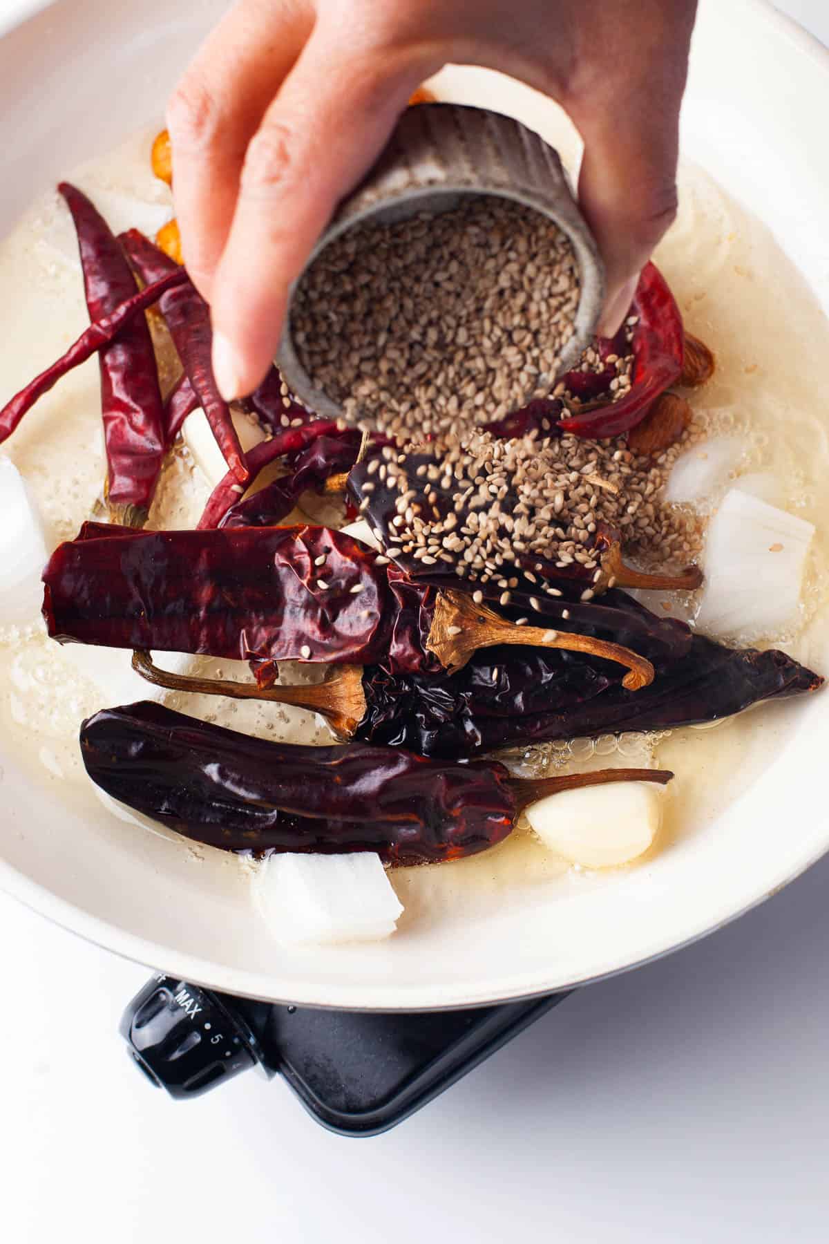 A hand pours sesame seeds into a white pan with dried red chilies, garlic cloves, onion chunks, and oil.