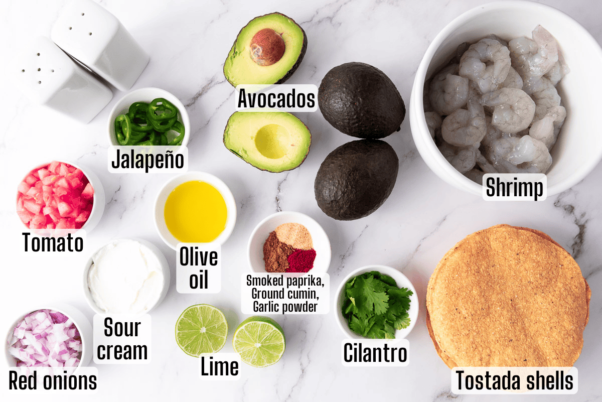 Labeled ingredients for shrimp tostadas, including shrimp, avocados, tostada shells, lime, cilantro, jalapeño, tomato, red onions, sour cream, olive oil, and spices on a white surface.